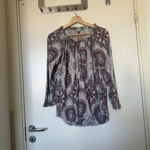 Odd molly blus  - Säljer en otroligt vacker blus i en äggvit färg med ett mönster i färgerna röd, rosa lila och grått. Från märket Odd Molly i storlek 2 som enligt deras storleksguide motsvarar en storlek M. Mycket fint skick, jag hittar inga synlig anmärkningar eller defekter.  