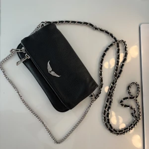 Svart axelväska Zadig & Voltaire - Säljer en svart axelväska i skinn från Zadig & Voltaire med silverfärgad kedjerem och coolt vingemblem framtill. Köpt tidigare här på Plick och den är fortfarande i mycket bra skick. Kan vara en aning rostig på få ställen vid kedjan