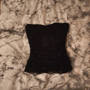 Svart spets tubtopp - Säljer en svart tubtopp i spets med blommigt mönster. Toppen är axelbandslös och har en tight passform som framhäver siluetten. Perfekt för dig som vill ha en stilren men ändå edgy look.