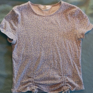 Ljusblå blommig topp från H&M - Supersöt ljusblå topp från H&M med små vita blommor över hela tyget. Modellen är kortärmad och har snygga snörningsdetaljer i sidorna nertill. Mjuk och stretchig bomullskänsla, perfekt för dig som gillar en söt och avslappnad stil. Storleken är 164 men funkar nog som en XS eftersom materialet är strechigt