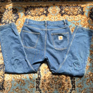Carhartt jeans  - Ett par klassiska blå jeans från Carhartt, modell Newel Pant. Är mestadels sydda i bomull med en liten del polyester för extra hållbarhet. Carhartt-logga på bakfickan och metallknapp framtill.