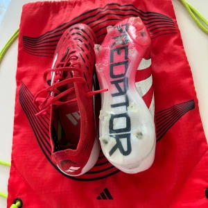 Adidas Predator fotbollsskor röd/vit - Säljer ett par Adidas Predator fotbollsskor i rött och vitt med klassiska tre ränder och snörning. Skorna har mönstrad ovandel och dobbar för bästa grepp på planen. Perfekta för dig som vill sticka ut med stil och prestanda på fotbollsplanen.