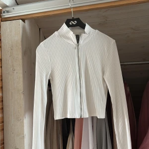 Vit ribbad croppad kofta med dragkedja - Säljer en vit croppad kofta från Divided med ribbad struktur och dragkedja framtill. Tröjan har lång ärm och en lite högre krage, perfekt att styla med jeans eller kjol för en clean look.