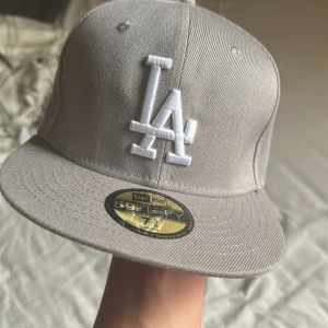 Grå LA Dodgers keps från New Era - Snygg grå 59FIFTY keps från New Era med LA Dodgers brodyr framtill i vitt. På sidan finns broderad 1988 World Series-logga och MLB-logga bak. Klassisk flat peak och stilren look, perfekt för dig som gillar streetwear och sport.