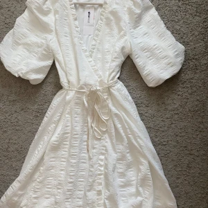 Vit omlottklänning från Gina Tricot - Superfin vit omlottklänning från Gina Tricot med puffiga korta ärmar, volangkant längs v-ringningen och knytband i midjan. Klänningen har en lätt struktur i tyget och är perfekt för sommarens alla tillfällen.