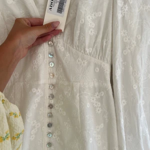 Vit blommig långklänning från Adoore - Superfin vit långklänning från Adoore med broderade blommor, v-ringning och långa puffärmar. Klänningen har knappar framtill och markerad midja för en snygg siluett. Perfekt för sommarens alla tillfällen!