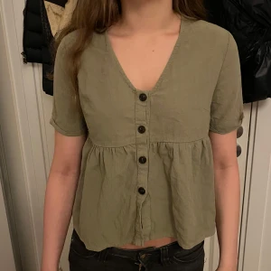 Olivgrön topp med knappar - Supersöt olivgrön blus med v-ringning och fyra svarta knappar framtill. Modellen är lite lösare med korta ärmar och lätt rynkad midja som ger en avslappnad och trendig look. Perfekt att styla med jeans eller kjol.