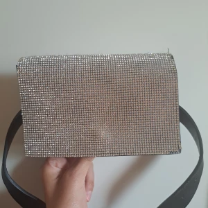 Glittrig silver midjeväska med strass - Säljer en snygg medjeväska med glittrig framsida täckt av strass i silver. Väskan har svart baksida och axelrem i svart syntetmaterial. Perfekt för dig som vill sticka ut och addera lite extra bling till din outfit.