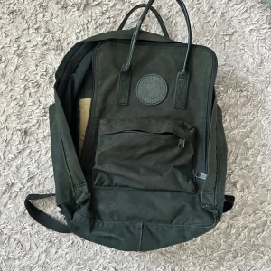 Svart Kånken ryggsäck från Fjällräven - Klassisk svart Kånken ryggsäck från Fjällräven med ikonisk rund logga framtill. Ryggsäcken har ett stort huvudfack med dragkedja, vadderat innerfack och en mindre ficka bak (perfekt att förvara datorn i). Tillverkad i slitstark polyester och bomull. Perfekt för skola eller cityhäng. Tillkommer en sittunderlägg, se bilden. Nypris 1295 kr