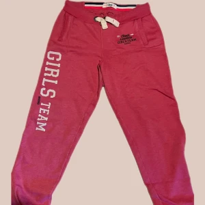 Rosa mjukisbyxor med tryck och snörning - Rosa mjukisbyxor med texten 'GIRLS TEAM' tryckt längs ena benet och snörning i midjan. Byxorna har ribbade muddar vid bensluten, fickor fram och en bakficka. Perfekta för chill eller träning.