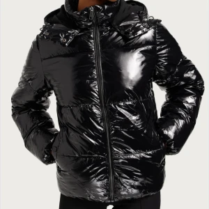 Svart glansig pufferjacka med huva - Säljer en svart, glansig pufferjacka med dragkedja och stor huva. Jackan har en oversized vibe, quiltad design och är perfekt för kalla dagar. Tillverkad i syntetmaterial och har långa ärmar. Snygg och trendig look som sticker ut.