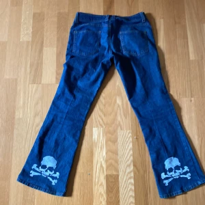 Blå bootcut jeans med döskalletryck - Säljer ett par blå jeans med bootcut-modell och coola vita döskalletryck längst ner på båda benen. Klassisk femficksdesign och normal passform. Perfekta för dig som vill sticka ut lite extra med din stil.