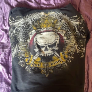 Svart hoodie Xzavier med dödskalletryck - Svart hoodie från Xzavier med stort dödskalletryck, vingar och krona i vitt, gult och rött. Texten 'hail the king' framtill. Dragkedja, huva och två fickor. 100% bomull, perfekt för dig som gillar edgy streetstyle.