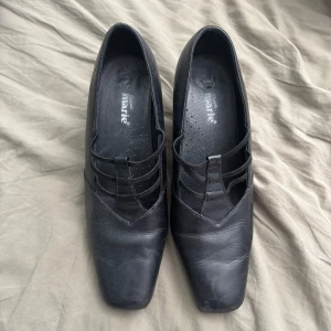 Svarta pumps med fyrkantig tå från Marie - Stilrena svarta pumps från Marie med fyrkantig tå och låg klack. Skorna har snygga remdetaljer över vristen och är tillverkade i skinn. Perfekta för dig som gillar en klassisk men edgy look.