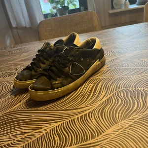 Svarta sneakers från Philippe Model - Snygga svarta sneakers från Philippe Model med vita detaljer och klassisk låg profil. Skorna har svarta skosnören, vit sula och är tillverkade i skinn. Ikonisk sköld på sidan och bekväm vadderad häl. Perfekta för en cool och avslappnad streetstil.  Skick 6/10 därav priset