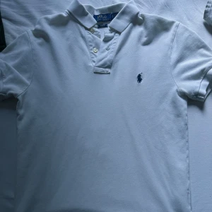 Vit pikétröja Polo Ralph Lauren - Klassisk vit pikétröja från Polo Ralph Lauren i slim fit-modell. Tröjan har korta ärmar, krage med knappar och den ikoniska broderade loggan på bröstet. Tillverkad i bomull för en skön och luftig känsla. Perfekt för en clean och stilren look.