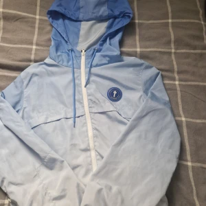 Trapstar blå vindjacka XS - Snygg ljusblå vindjacka från Trapstar med huva och vit dragkedja. Jackan har coolt Trapstar-tryck på ryggen och rund blå logga på bröstet. Tillverkad i lätt polyester, perfekt för vår och sommar. Regular fit och långa ärmar. Fräsch streetstil!