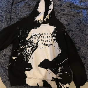 Maison Margiela svartvit hoodie - Säljer en Maison Margiela hoodie i svart och vitt med coolt abstrakt mönster och tryckt logga framtill. Tröjan har huva och långa ärmar, tillverkad i mjuk bomull. Perfekt för dig som gillar statement-plagg och streetstyle.