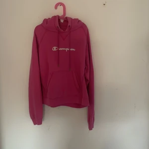 Rosa hoodie från Champion - Säljer en snygg rosa hoodie från Champion med vit logga på bröstet. Tröjan har huva med snörning och en stor magficka framtill. Perfekt för dig som gillar streetwear och vill ha en poppig färg i garderoben. Obs den ser lite mer skrik rosa/röd ut än vad den är i verkligheten