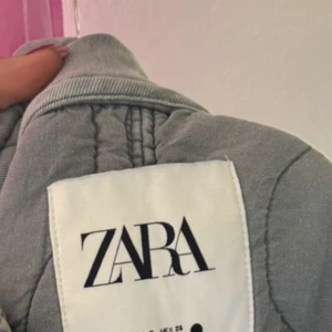Grå quiltad dunväst från Zara - Säljer en grå quiltad dunväst från Zara med hög krage och två stora fickor framtill. Västen har ett unikt mönster i sömmarna och stängs med dragkedja. Perfekt att slänga över en hoodie eller tröja för en chill look. Helt ny utan lapp