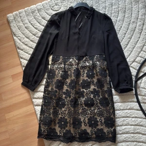 Svart kortklänning med blommig spets - Supersnygg svart kortklänning med långärmade ärmar och v-ringning. Kjoldelen har beige botten med svart blommig spets ovanpå som ger en cool kontrast. Överdelen är enfärgad och har en lätt rynkad detalj vid kragen. Perfekt för dig som gillar unika detaljer!
