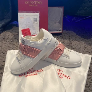 Valentino Garavani Rockstud sneakers - Snygga vita Valentino Garavani Rockstud sneakers med bred rosa rem över foten dekorerad med ikoniska nitar. Skorna har rund tå, platt sula och är tillverkade i skinn. Perfekta för dig som vill sticka ut med en lyxig och trendig look. Skorna är ej andvända, bara testade, säljs pga av fel storlek vid online beställning. Hör av er så svarar vi på era frågor! Priset är ej fast så skriv för ett rimligt pris! Varan är såklart äkta och kommer med äktighets bevis och kvitto✨