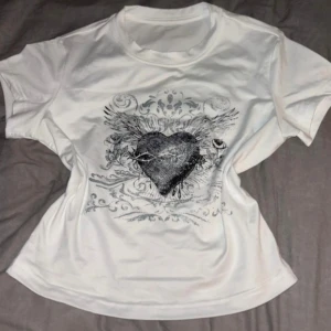 Vit t-shirt med hjärttryck (inte min bild) - Vit t-shirt med coolt svart hjärta och vingar tryckt på bröstet. T-shirten har rund halsringning och korta ärmar. Materialet känns mjukt och skönt, perfekt för en avslappnad stil.