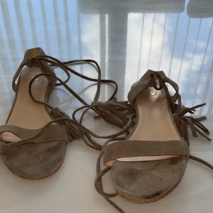 Beige mockasandaler med snörning - Snygga sandaler i beige mocka med platt sula och rund tå. De har långa snören som kan lindas runt vristen och avslutas med tofsar för en trendig boho-look. Perfekta för varma dagar och ger en avslappnad men stilren vibe.