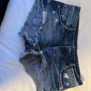Blå jeansshorts med slitningar från Zara - Snygga blå jeansshorts från Zara Trafaluc med råa, fransiga kanter och slitna detaljer. Shortsen har ett coolt paisleymönstrat blått tyg bakom de trasiga partierna och klassisk femficksmodell. Perfekta för sommaren och ger en avslappnad vibe.