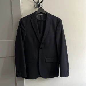 Svart kavaj H&M Essentials Slim Fit - Snygg svart kavaj från H&M Essentials, modell No 12 i slim fit. Klassisk design med två framfickor med lock, bröstficka och knappar vid ärmslut. Perfekt för dig som vill ha en stilren och modern look. Passar perfekt till jeans eller kostymer. Storlek 46, pris kan diskuteras. Endast använd en gång