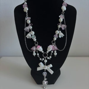 Sött halsband med pärlor och rosett - Unikt halsband med silverfärgad kedja, vita och rosa pärlor, blommor i plast och en stor vit rosett. Hängsmycke med måne och hjärta i silver och rosa. Perfekt statement-piece för dig som gillar gulliga detaljer och pastellfärger.