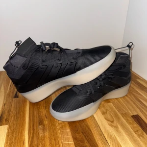 adidas x Fear of God Athletics I Carbon sneakers - Stilrena svarta sneakers från Adidas med högt skaft och tydliga svarta ränder på sidan. Skorna har snörning och en chunky, vit platt sula som ger en modern look. Ovandelen är i syntetmaterial med diskreta detaljer och sportig känsla.