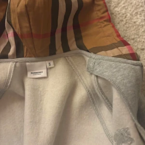 Grå zip-hoodie från Burberry S - Grå zip-hoodie från Burberry med klassiskt rutigt foder i huvan. Jackan har dragsko i huvan, dragkedja framtill och är tillverkad i mjuk bomullsmix. Perfekt för dig som gillar stilren design med en touch av lyx.