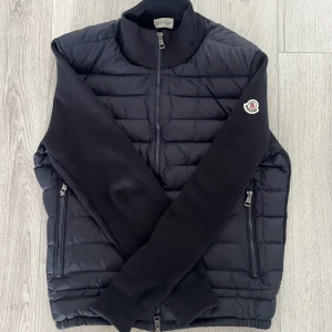 Svart Moncler dunjacka med stickade ärmar - Snygg svart dunjacka från Moncler med quiltad framsida och stickade ärmar. Perfekt för dig som vill ha en stilren statement-piece. Pris kan diskuteras vid snabb affär