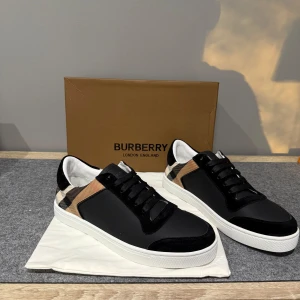 Burberry skor - Snygga sneakers från Burberry med klassiskt rutigt mönster i beige, svart och vitt på sidorna. Ovandelen är i svart skinn och mocka, med vita sulor och svarta snören. Perfekt för dig som vill ha en stilren och lyxig look med en touch av streetstyle.