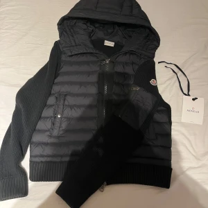 Svart Moncler moncler cardigan  - Snygg svart moncler cardigan från Moncler med stickade ärmar och quiltad kropp. Jackan har huva, dragkedja framtill och Moncler-logga på ärmen. Perfekt för dig som vill ha en stilren och exklusiv look under kyliga dagar.