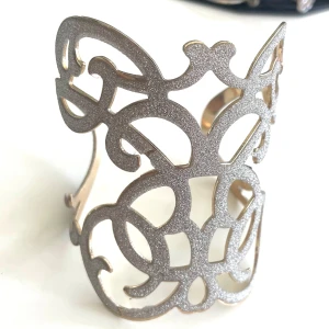 Stylish, gnistrande, justerbart armband/armring - Ett Superstylish och snyggt lite större armband som är mycket justerbart genom att klämma fast det på plats. Det är guldfärgat på insidan och ytan som en gnistrande matta. Mycket bra skick, har CC aldrig använt det. Mått  Höjd fram 8 cm H bak. Vid öppning 3,5 cm Omkrets 8 cm i diameter men justerbart Vikt 35 gram 