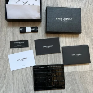 YSL krokodilmönstrat läder med svart text - Säljer en lyxig YSL (Saint Laurent) korthallare i svart präglat krokodilmönstrat läder med elegant silverfärgad logotyp. Tidlös och sofistikerad design med plats för flera kort. Akta designeraccessoar som kombinerar stil och funktion. i mycket fint skick.