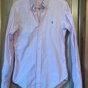 Rosa rutig skjorta Polo Ralph Lauren - Säljer en rosa och vit rutig skjorta från Polo Ralph Lauren i slim fit. Skjortan har lång ärm, klassisk krage och knappar framtill. Broderad blå logga på bröstet. Perfekt för dig som gillar stilrena och klassiska plagg.