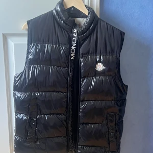 Svart dunväst från Moncler - Säljer en svart, glansig dunväst från Moncler med hög krage och tydlig logga på bröstet. Västen har dragkedja framtill med Moncler-tryck och två fickor med knappstängning. Perfekt för lager-på-lager och riktigt snygg till streetwear.