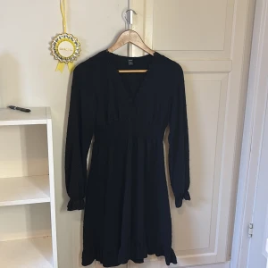 Svart kortklänning med spets från SHEIN - Svart kortklänning från SHEIN med lång ärm, v-ringning med spetskant och smockad midja. Tunt tyg med diskret prickigt mönster och volangdetalj nertill. Perfekt för dig som gillar en feminin och stilren look.