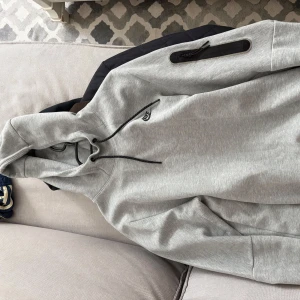 Grå nike tech baggy dress - Snygg ljusgrå dress från Nike med svart snörning i huvan och diskret Nike-logga på bröstet. Tröjan har lång ärm, praktisk dragkedja på ena ärmen och är tillverkad i mjuk bomullsblandning. Perfekt för en sportig och avslappnad stil.