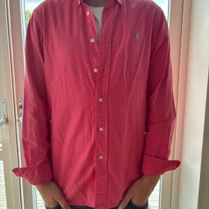 Rosa skjorta från Polo Ralph Lauren - Snygg rosa skjorta från Polo Ralph Lauren i bomull. Klassisk modell med knappar hela vägen, krage och broderad blå logga på bröstet. Långärmad och passar perfekt till jeans eller chinos. Skön och stilren vibe.