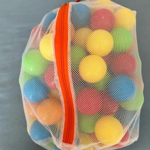 Vit nätväska med orange dragkedja - Vit nätväska med orange dragkedja och handtag. Väskan är genomskinlig och har en fyrkantig form, perfekt för att bära leksaker eller sportprylar. Tillverkad i syntetmaterial och stängs med dragkedja längs framsidan.