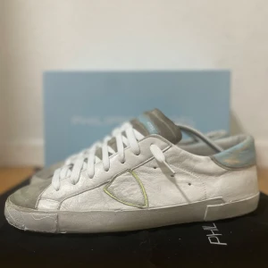 Vita sneakers från Philippe Model - Snygga vita sneakers från Philippe Model med grå detaljer och ljusblå häl. Skorna har klassisk snörning, rund tå och är tillverkade i skinn. En gul kontrastsöm runt logotypen på sidan ger en cool touch. Perfekta för en avslappnad streetstyle. Storlek 44, bra skick! Nypris runt 3000 kr. Allt org medföljer.