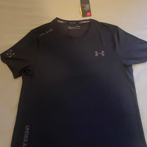Svart Under Armour tränings-tshirt - Svart t-shirt från Under Armour med diskret logga på bröstet och tryck på ärmen och sidan. Tillverkad i ett lätt och snabbtorkande syntetmaterial, perfekt för träning. Klassisk rund hals och korta ärmar för en sportig look. lyser i mörkret. Oanvänd 