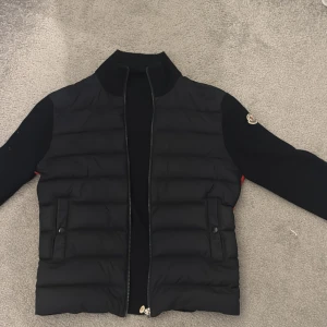 Svart Moncler cardigan - Snygg svart pufferjacka från Moncler med quiltad front, långa ärmar och dragkedja. Röda ränder och Moncler-logga längs sidorna ger en sportig vibe. Jackan har två fickor med knappar och klassisk Moncler-patch på ärmen. Perfekt för dig som gillar streetstyle.