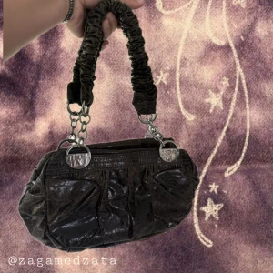 Brun handväska från Miss Sixty - Cool brun handväska från Miss Sixty i glansigt skinnliknande material. Väskan har scrunchade handtag med silverfärgade kedjor och logga, samt dragkedja upptill. Insidan är fodrad med leopardmönster och har flera fack. Perfekt för dig som gillar unika detaljer.