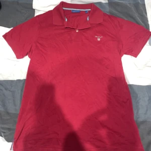 Röd pikétröja från GANT, storlek S - Snygg röd pikétröja från GANT i klassisk modell med krage och korta ärmar. Tröjan har broderad GANT-logga på bröstet och är gjord i mjuk bomull. Perfekt för en stilren och avslappnad look.