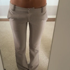 Beiga kostymbyxor med bootcut - Snygga beiga kostymbyxor med bootcut-snitt och hög midja. Byxorna har bälteshällor, diskreta bakfickor och stängs med dragkedja och knapp. Perfekta för en clean och stilren look.